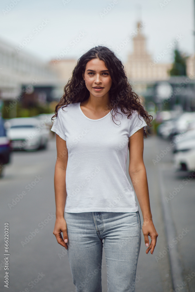 Attractive brunette woman with curly hair in a white t-shirt walks on the street.