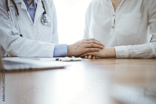 Doctor and patient sitting near each other at the wooden table in clinic. Female physician's hands reassuring woman. Medicine concept
