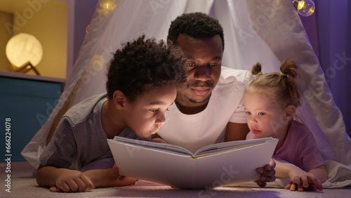 African American father reads bedtime stories to preschooler boy and toddler girl.