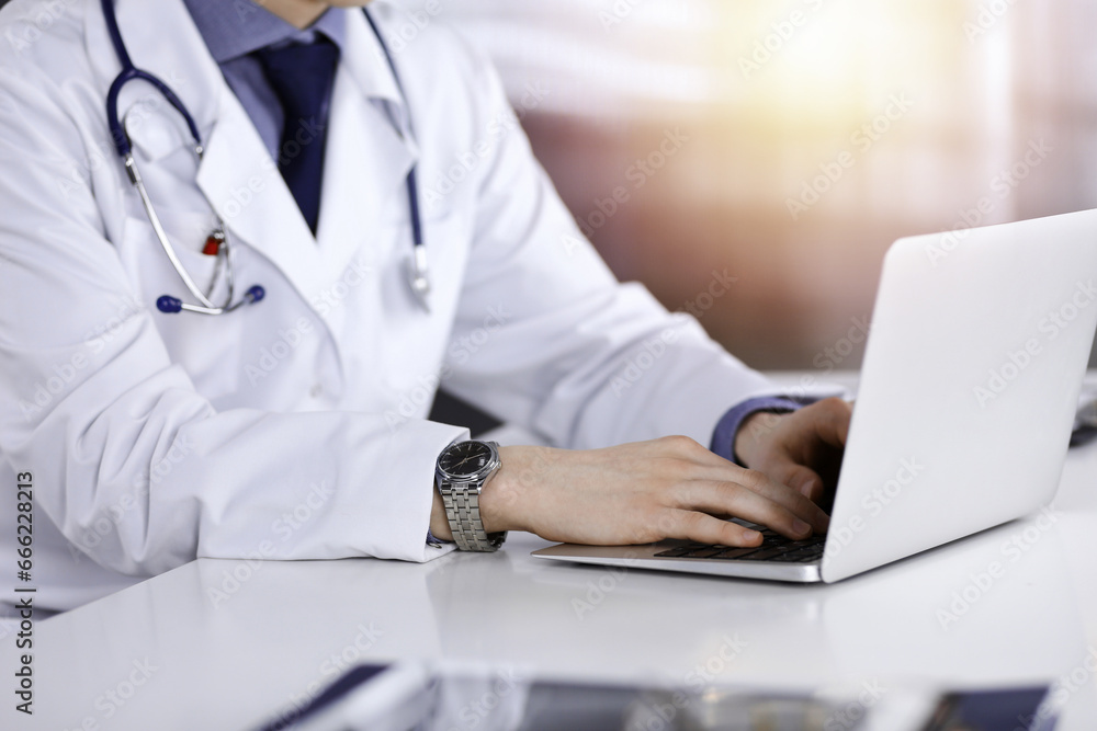 Unknown male doctor sitting and working with laptop in a darkened clinic, glare of light on the background, close-upof hands Unknown male doctor sitting and working with laptop in a darkened clinic, glare of light on the background, close-upof hands