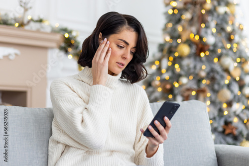 Upset woman sitting alone at home on sofa in living room near Christmas tree, Woman celebrating new year and Christmas, holding phone, received online notification with bad news, unhappy reading