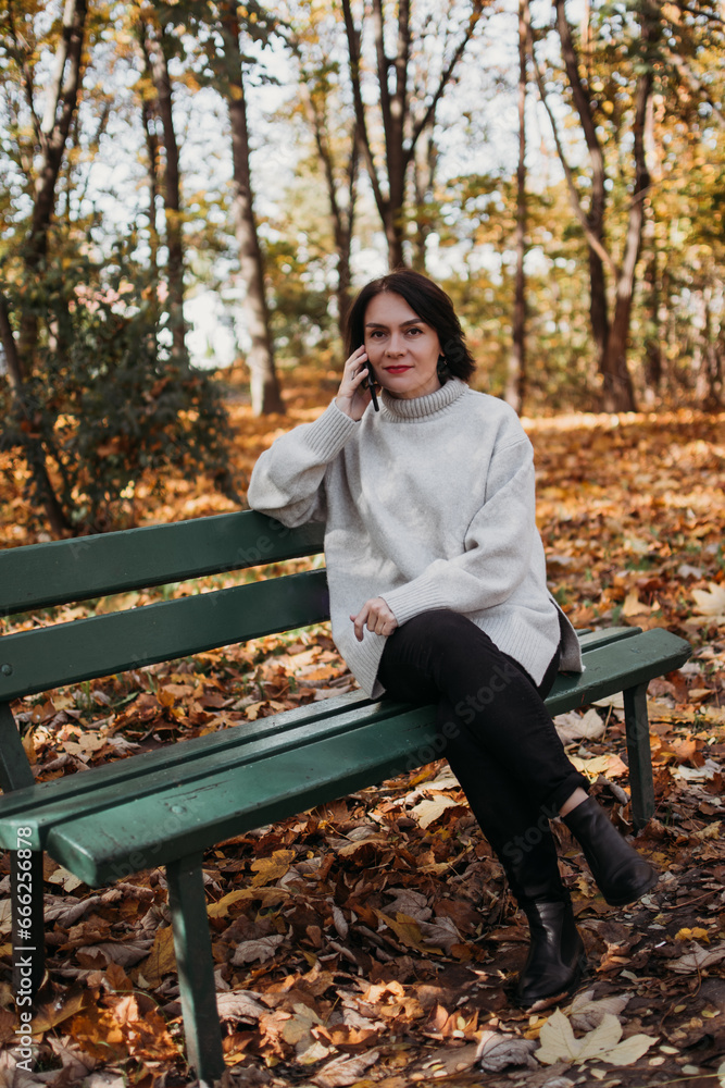Smiling woman sitting on bench in autumn park using phone Smiling woman sitting on bench in autumn park using phone