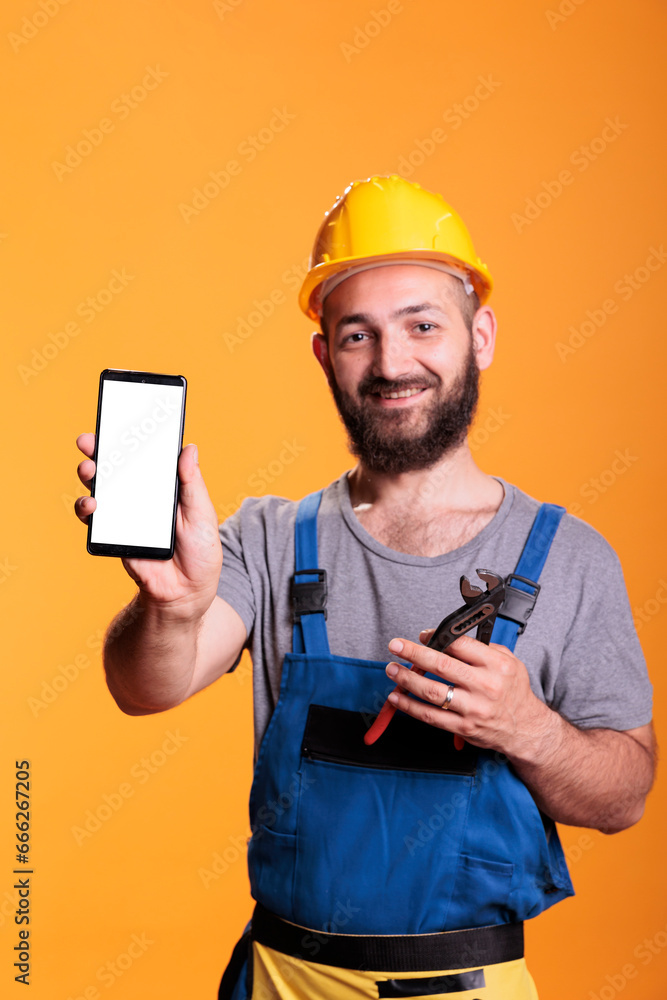 Construction worker holding cell phone with empty screen for advertising in front of camera. Professional builder with hard hat against yellow background in studio shot. Construction worker holding cell phone with empty screen for advertising in front of camera. Professional builder with hard hat against yellow background in studio shot.