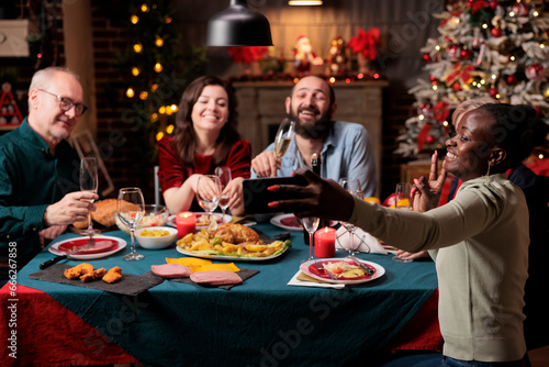 People taking pictures at dinner table  making memories of christmas eve festivity gathered around at home. Diverse persons having fun with wine and food  taking photos with smartphone device.