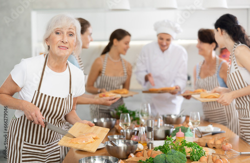 Mature woman holding cutting board with raw chicken breast in her hands posing surrounded by other members of cooking course