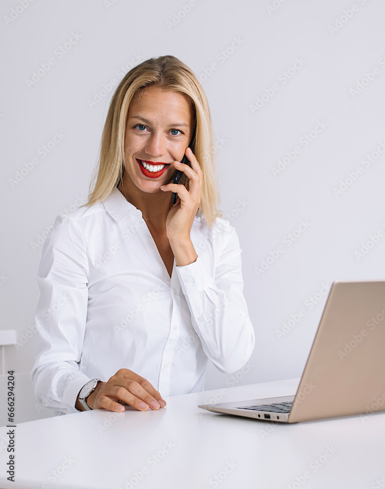 Glad female sitting at table talking on mobile and working on laptop Glad female sitting at table talking on mobile and working on laptop