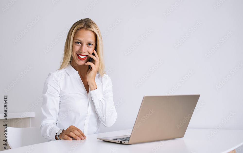 Glad female sitting at table talking on mobile and working on laptop Glad female sitting at table talking on mobile and working on laptop