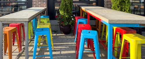 Colorful iron metal stackable bar stools, blue, red, orange, and yellow in color. The cafe chairs are next to rows of long wooden pub tables on the outside of a restaurant on the patio garden.