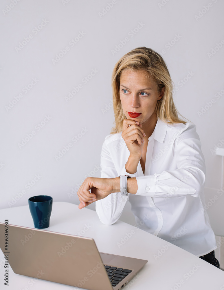 Young thoughtful blonde checking time and sitting at desk with laptop Young thoughtful blonde checking time and sitting at desk with laptop