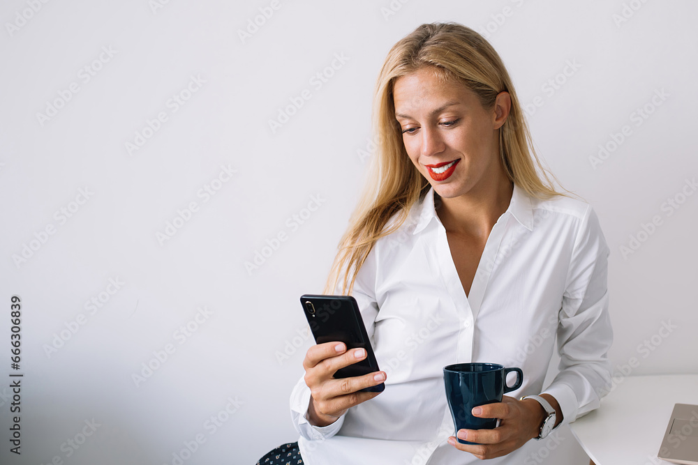 Serious female sitting at table with blue mug messaging on phone and working on laptop Serious female sitting at table with blue mug messaging on phone and working on laptop
