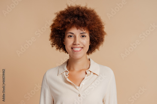 Portrait of beautiful smiling curly haired African American woman wearing stylish shirt looking at camera isolated on beige background. Natural beauty concept © Maria Vitkovska