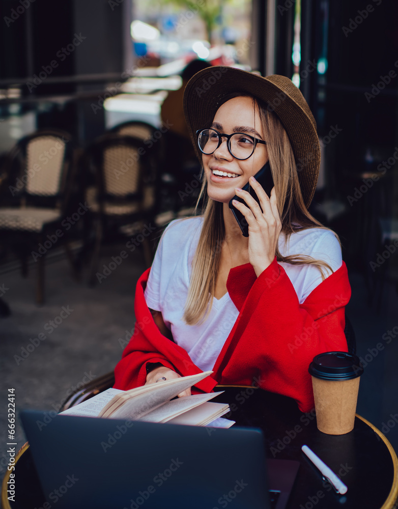 Fototapeta premium Joyful hipster woman in hat talking on smartphone while working at table at cold cafe terrace