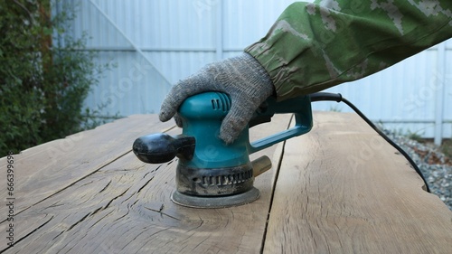 carpenter in gloves works with a sander in a green case with a round emery attachment on polishing solid wooden oak boards in an outdoor workshop, smoothing the surface of the wood with emery photo