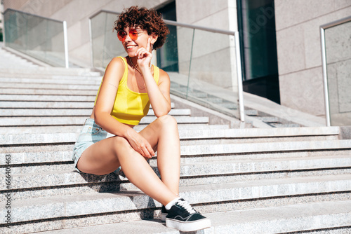 Young beautiful smiling hipster woman in trendy summer clothes. Carefree woman with curls hairstyle, posing in street. Positive model outdoors. Cheerful and happy, sits at stairs. In sunglasses