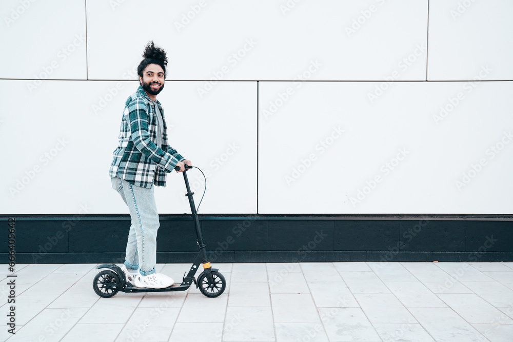Trendy smiling bearded man in casual clothes riding electric scooter in urban background. Handsome model posing in the street near wall. Hipster guy with curly hairstyle. Cheerful and happy Trendy smiling bearded man in casual clothes riding electric scooter in urban background. Handsome model posing in the street near wall. Hipster guy with curly hairstyle. Cheerful and happy