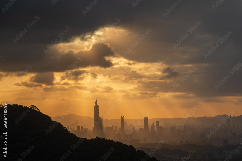 At dusk, the sun breaks through the clouds and shines on Taipei City. Silhouettes of city buildings. Orange sky and dark clouds.