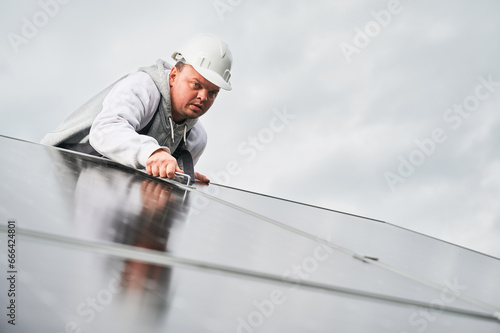 Man roofer mounting photovoltaic solar panels on roof of house. Engineer in helmet installing solar module system with help of hex key. Concept of alternative, renewable energy.