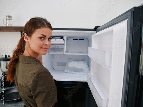 Woman smiling with teeth looking into camera in kitchen at home opened freezer empty with ice inside, home refrigerator, defrosted, view from back, stylish interior.