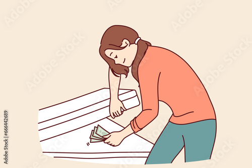 Woman hides money under mattress and smiles, keeping money safe and wanting to save up for big purchase. Concept of lack of financial literacy in girl who does not trust bank deposits