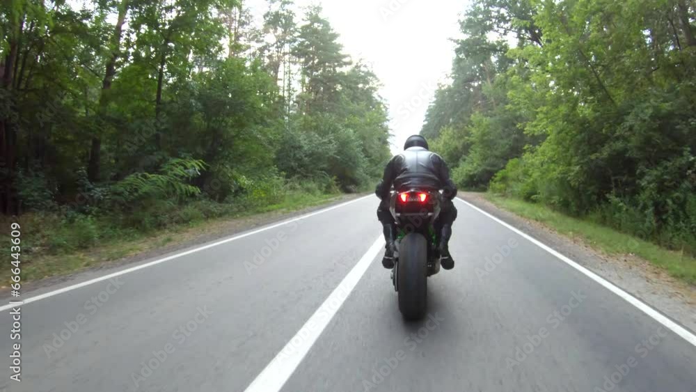 Follow to motorcyclist rides on motorcycle on woodland country road. Man in helmet races modern sport motorbike at rural route near forest. Guy driving bike during trip or journey. Concept of freedom Follow to motorcyclist rides on motorcycle on woodland country road. Man in helmet races modern sport motorbike at rural route near forest. Guy driving bike during trip or journey. Concept of freedom