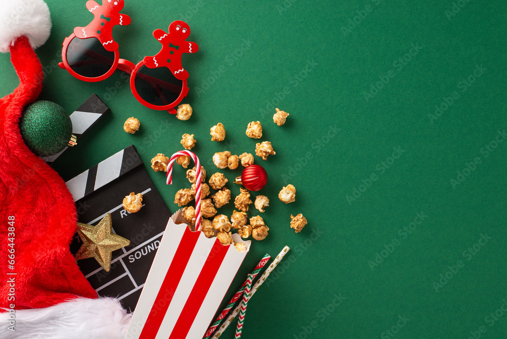 Christmas-themed premiere featuring thematic elements. Top-view of movie clapper, gingerbread man-shaped glasses, delectable popcorn, Santa's hat, ornaments, straws on green backdrop with ad space