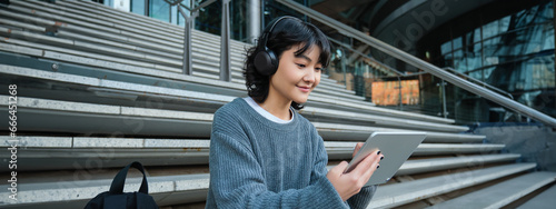 Smiling chinese student sits on stairs with tablet, draws digital art, graphic design project for freelance job, listens music in headphones and smiles