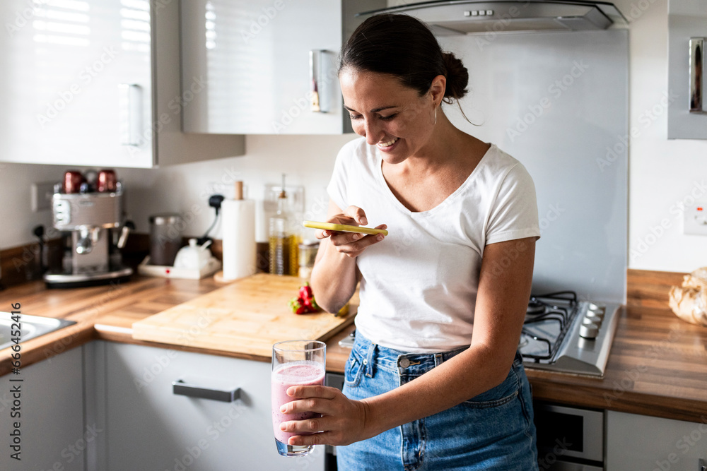 Happy woman photographing milkshake through smart phone in kitchen at home Happy woman photographing milkshake through smart phone in kitchen at home