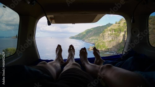 Young traveler couple laying in camping trailer bed, enjoying view from open window and swinging their feet. Cinematic view from cozy camping trailer to mountains and ocean. Travel by camping trailer.
