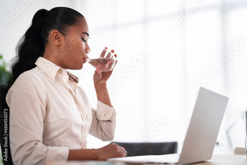 Side view of young Vietnamese female employee in formal clothes drinking refreshing water from glass while sitting at table with computer in office photo