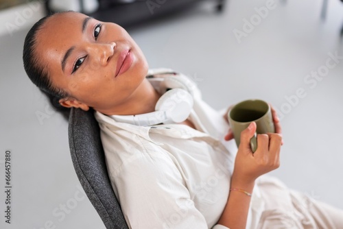 High angle of young female entrepreneur in glasses with headphones around neck sitting at workplace and drinking hot beverage from cup during break at work photo