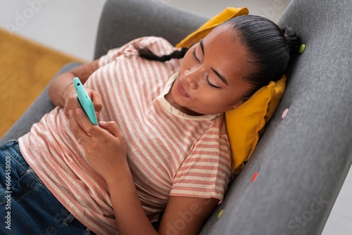 Young female Vietnamese in casual outfit lying on couch and messaging via mobile phone during break in modern apartment photo