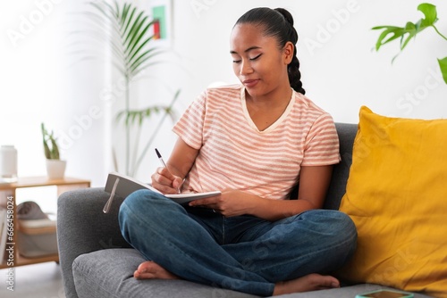 Young Asian female sitting on sofa wearing striped t shirt writing on notepad with pen during free time photo