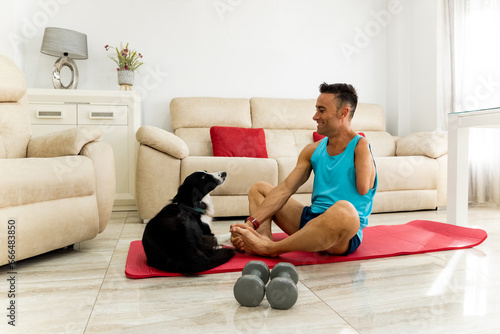 An adult man with an amputated arm exercises in his living room with his border collie dog. The athlete is stretching his legs while he looks at his dog. Concept of healthy living at home.
