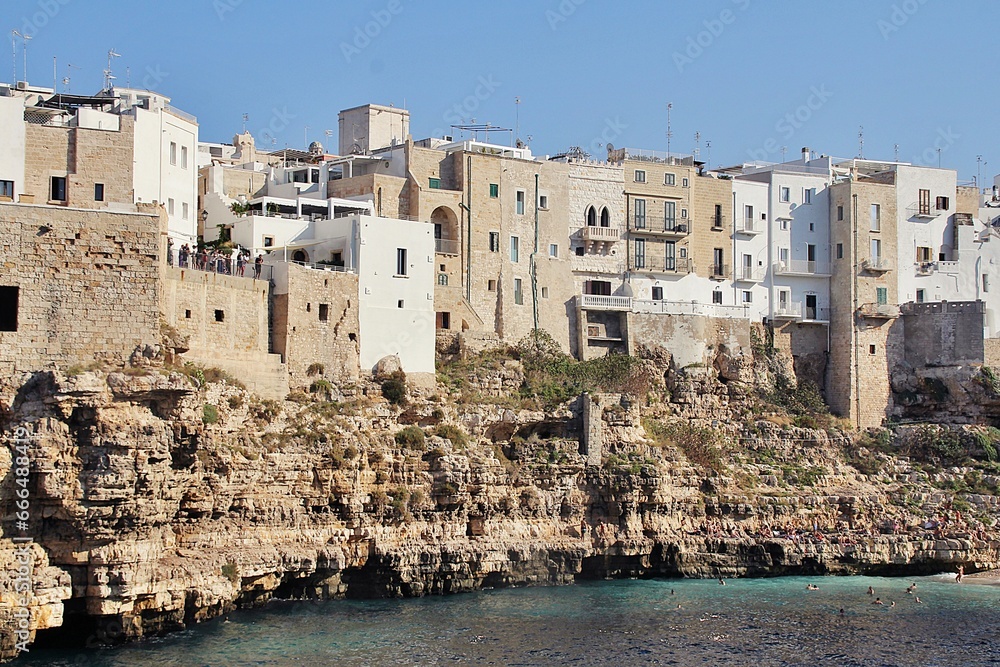  large cliff on the seashore, rocks by the sea, buildings built on the rocks right on the seashore, Polignano a Mare, rocky coast, turquoise water, people bathing in the sea
