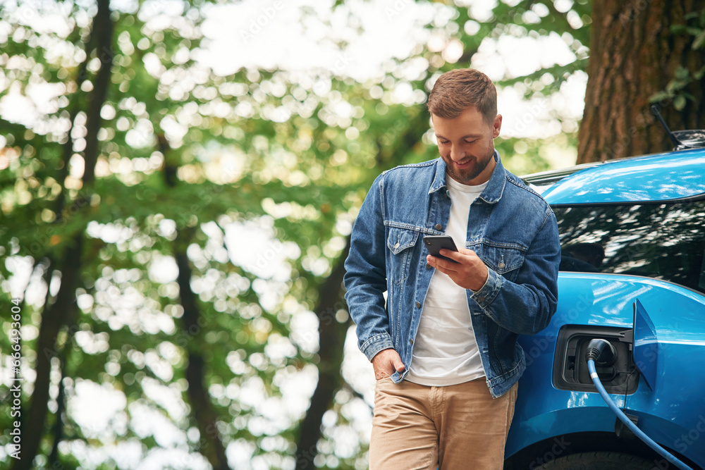 Fototapeta premium Standing and holding smartphone. Handsome man is waiting for his electric car to charge, outdoors