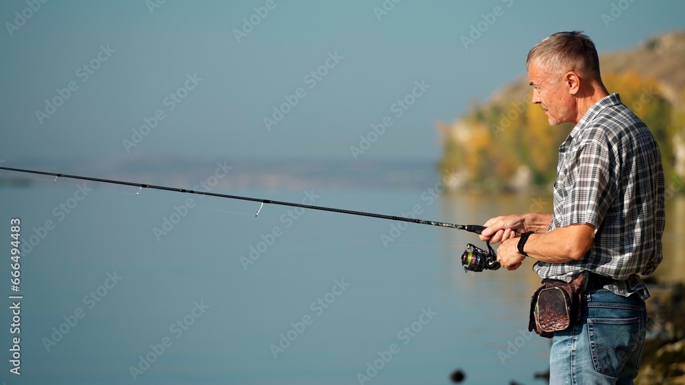 In autumn, a mature adult man in a shirt and jeans catches fish with a spinning rod on the river bank. Senior adult goes fishing on weekends. A young pensioner is engaged in a hobby. In autumn, a mature adult man in a shirt and jeans catches fish with a spinning rod on the river bank. Senior adult goes fishing on weekends. A young pensioner is engaged in a hobby.