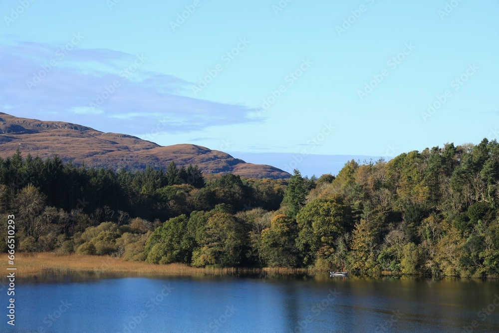 Landscape at Lough Gill, County Leitrim, Ireland on autumn day featuring small boat close to shore against backdrop of forest, Killery Mountain and blue sky