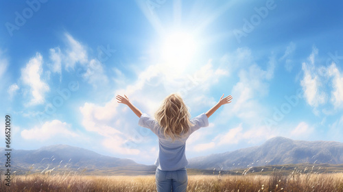 A beautiful blonde hair woman with her arms raised in the air on a blue sky day with a beautiful landscape behind her