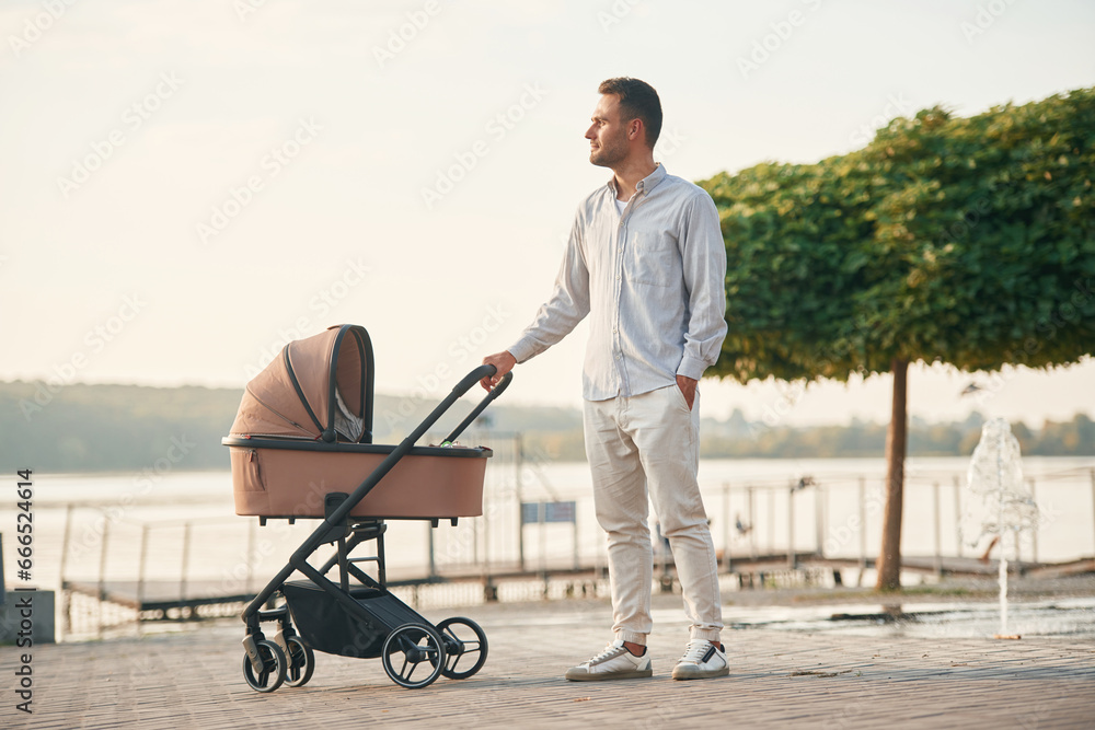 Green tree behind. Young father is outdoors, standing with pram