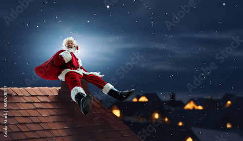 Cheerful, happy senior man Santa Claus sitting on chimney on roof at night with big bag with presents. Concept of winter season, holidays, fantasy, joy and fun, Christmas