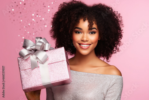 Beautiful african american girl holding gift box in her hands. Pretty curly hair woman posing with xmas present in her arms, pink studio background photo