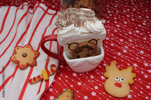 A Santa Claus-shaped mug with cream, stained with cocoa powder. A chocolate chip cookie peeks out of the cup. Homemade Christmas cookies. Red tablecloth with white stars and a striped kitchen towel.  photo