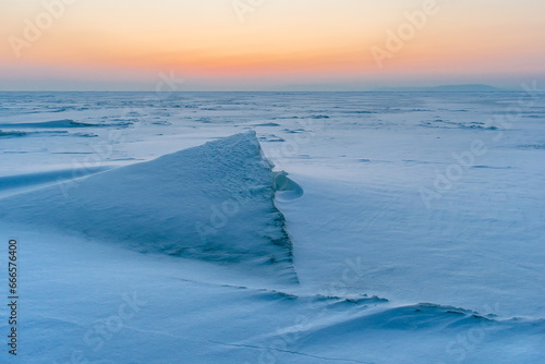 Snowdrifts on the ice surface during sunset. Winter landscape. A frozen surface of ice with snow intersected by many cracks. © Tishina