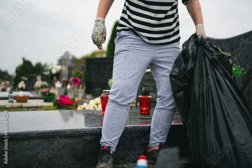 Professional gravesite care. Big autumn cemetery cleaning before the All Saints Day. Unrecognizable worker with big plastic bag and protective gloves collecting garbage.