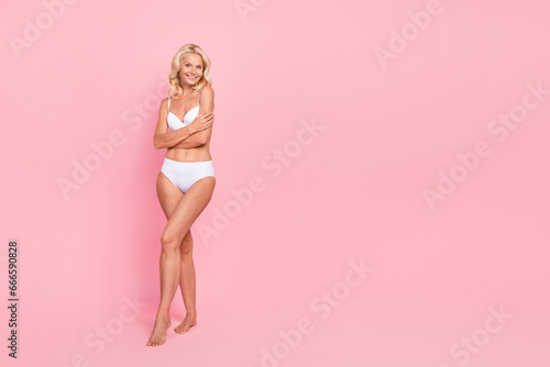 Full size photo of attractive elderly woman embrace shoulders posing wear trendy white lingerie isolated on pink color background