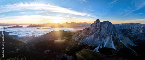 Fototapeta Naklejka Na Ścianę i Meble -  Sunrise over the Sass de Puta mountain peak at Passo delle Erbe pass against the Dolomite peaks in the background, inverse cloud cover in the valley, sun rays. Aerial drone mountainscape panorama.