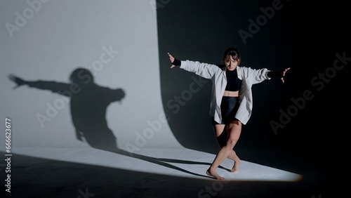 Young woman wearing a top, shorts and a shirt performing contemporary dance under a spotlight in the studio. Contrasting black falling shadow on the background. Full length.