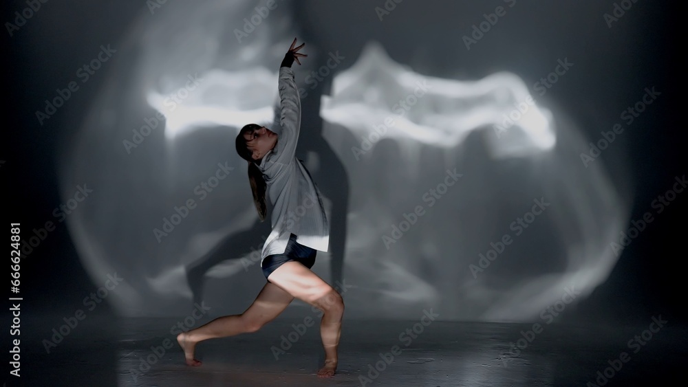 Young woman wearing a top, shorts and a shirt performing contemporary dance under the glare of the light in the studio. Contrasting black falling shadow on the background. Full length.