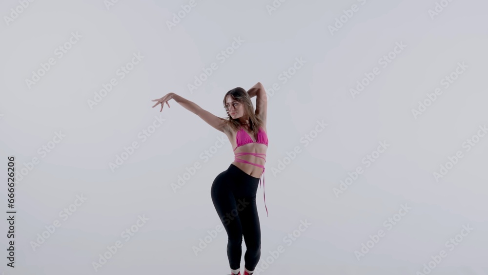 Young woman wearing a bright pink bra, top, leggings and high heels performing energetic heels dance or posing in studio, isolated white background. Full length