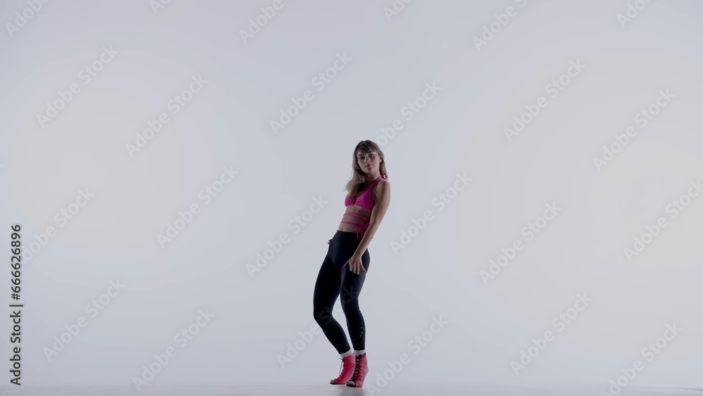 Young woman wearing a bright pink bra, top, leggings and high heels performing energetic heels dance or posing in studio, isolated white background. Full length